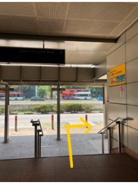 The sheltered MRT exit at street level: a yellow arrow curves right towards the handrail and the road beyond.