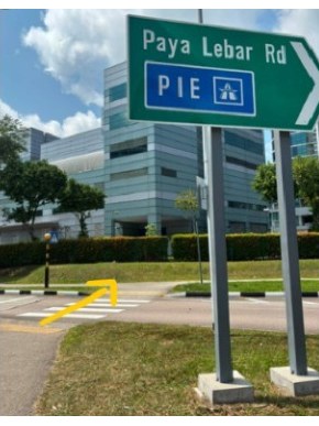 A green Paya Lebar Road and PIE direction sign beside the pavement, with a yellow arrow pointing along the zebra crossing towards the bus stop.